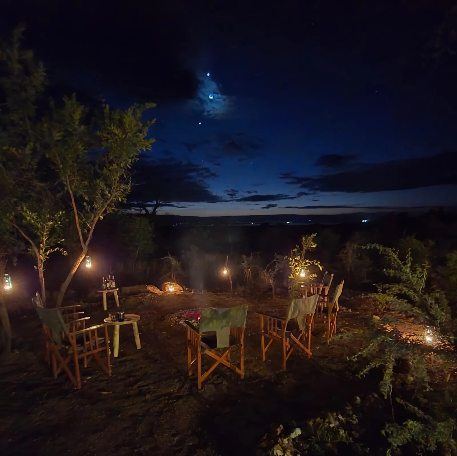 Campfire lounge at night with lanterns in the trees, the moon above Lake Burunge