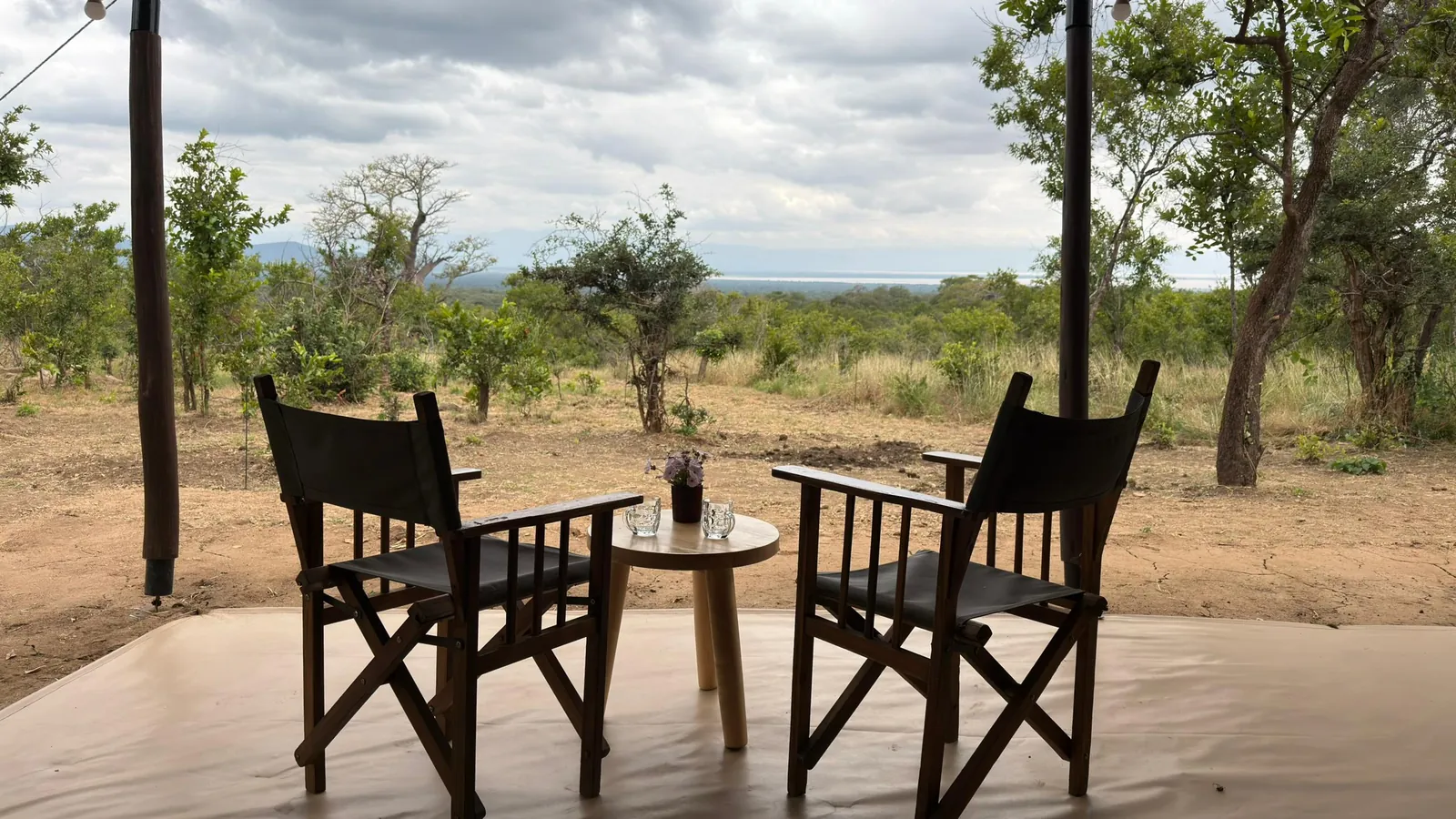 Two safari chairs and a small table on a tent veranda, looking out over the bush to Lake Burunge